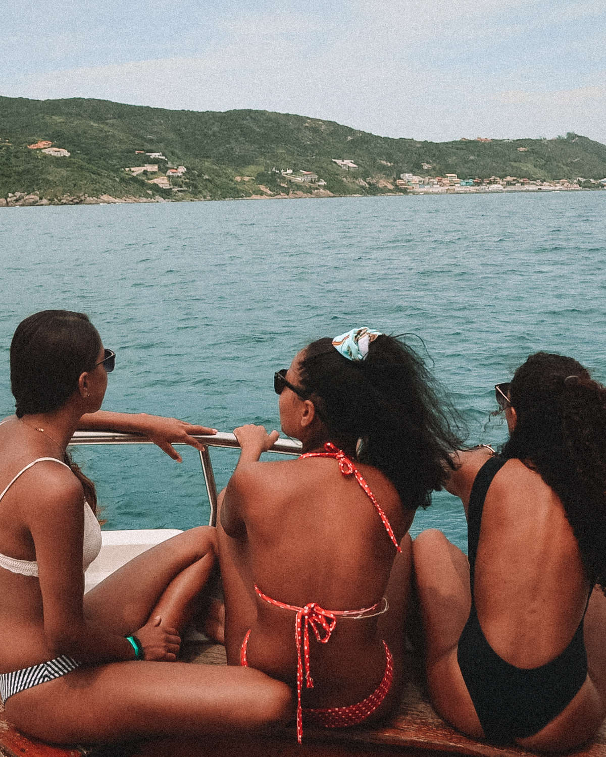 Three women enjoying a boat tour in Arraial do Cabo, sitting at the back of the boat and taking in the coastal scenery. The green mountains and scattered houses on the hills are visible in the distance, highlighting a relaxing and scenic activity in this popular tourist destination.