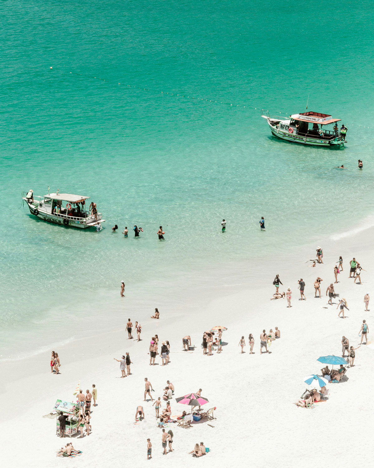 Aerial view of a lively scene at **Prainhas** do Pontal do Atalaia in March, the best time to visit Arraial do Cabo, featuring crystal-clear turquoise water, with scattered people, swimmers, and two boats near the shore.