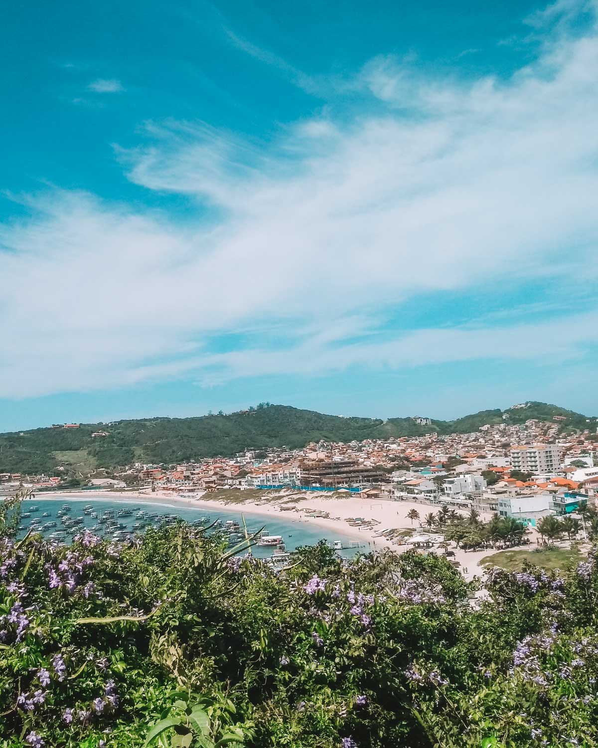 View of Arraial do Cabo featuring a bay full of boats at Playa dos Anjos, showcasing where to stay in Arraial do Cabo. The scene includes a sandy beach and a town sprawling across hills under a blue sky with thin clouds. Purple flowers beautifully frame the foreground of the image.