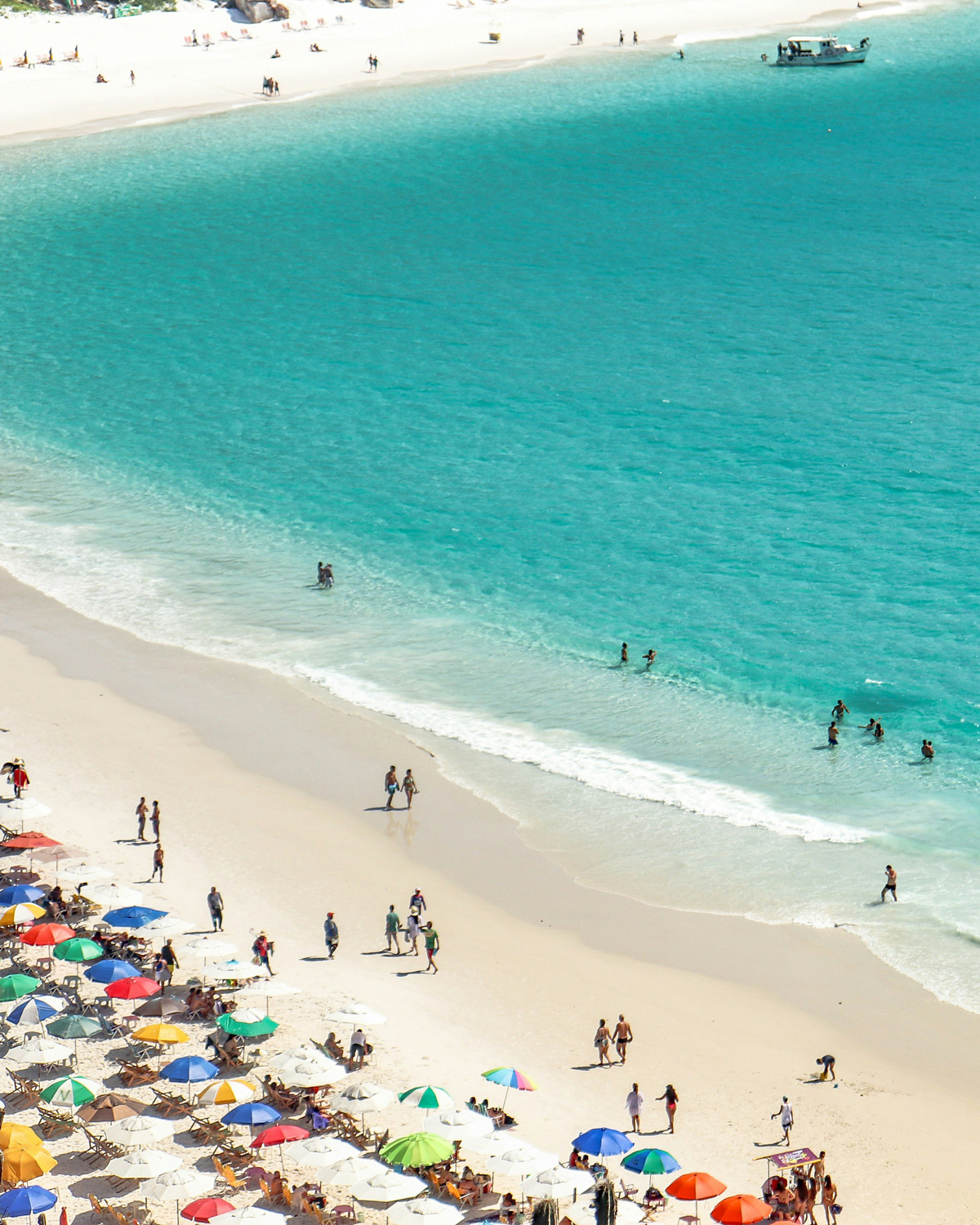 Aerial view of **Prainhas do Pontal do Atalaia** in Arraial do Cabo, showing tourists relaxing under colorful umbrellas and swimming in crystal-clear waters, perfect for those looking for things to do in this tropical destination.