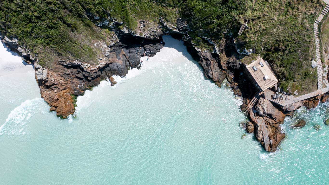 Aerial view of the rocky coastline with turquoise water and a small building on the cliff at Praianhas do Pontal do Atalaia in Arraial do Cabo. The waves gently break against the rocks, and a path leads to the structure.