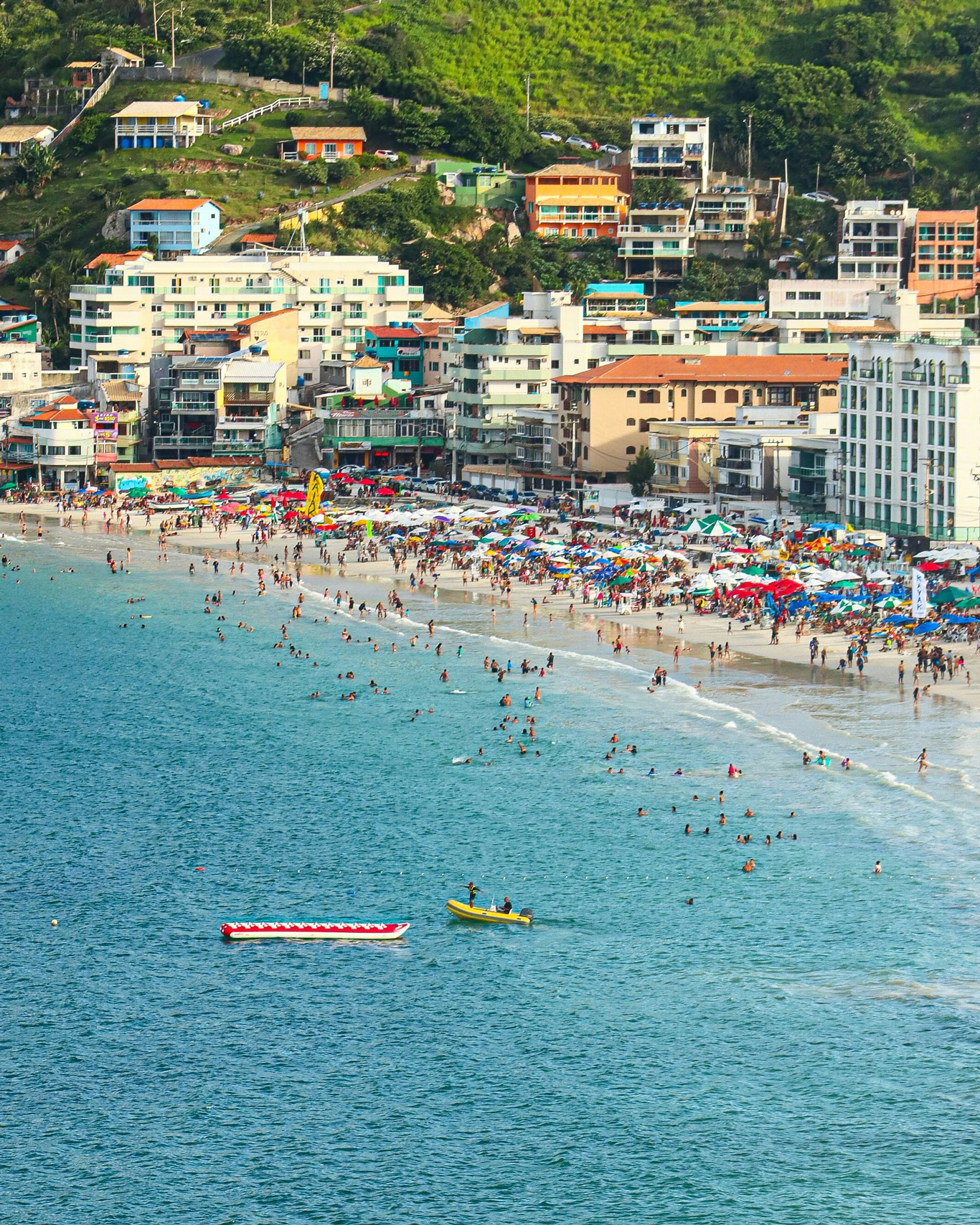 Aerial view of Prainha during summer, a bustling beach in Arraial do Cabo, filled with numerous beachgoers and colorful umbrellas dotting the sand. The beach is encircled by multi-story residential buildings and green hills in the background, with a couple of boats in the sparkling blue water.