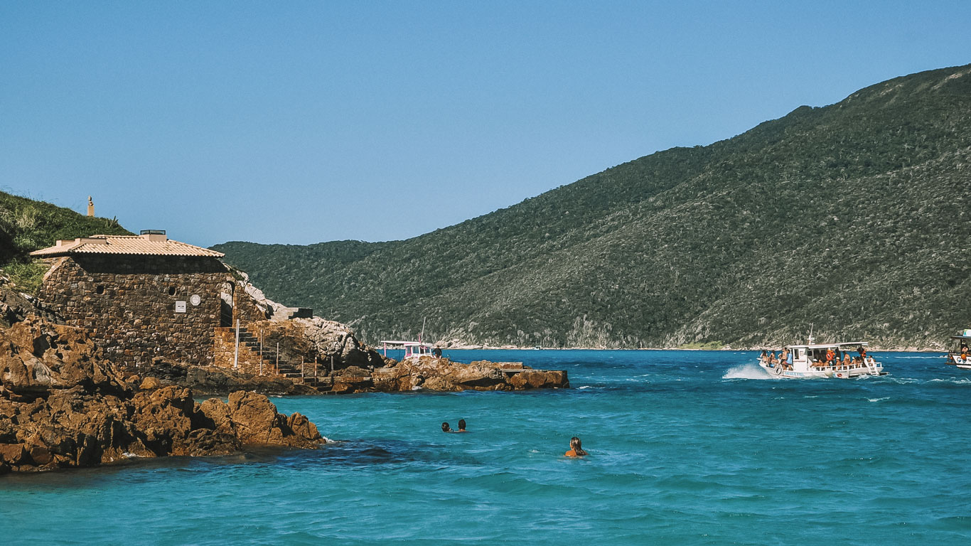 Photograph of a beach in Arraial do Cabo with crystal clear turquoise water and sunbathers enjoying the day. On the left, a stone structure with a thatched roof is in front of hills covered in vegetation, while a boat full of tourists sails to the right.