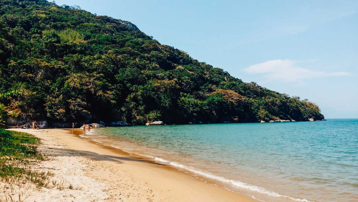 View of Praia do Pouso, one of the most peaceful beaches to stay at in Ilha Grande. This beach, located in Ilha Grande, Rio de Janeiro, is known for its soft, light sand blending with crystal-clear blue waters. A dense green forest covers the hills to the left, while a few people stroll or relax near the water on a sunny day.