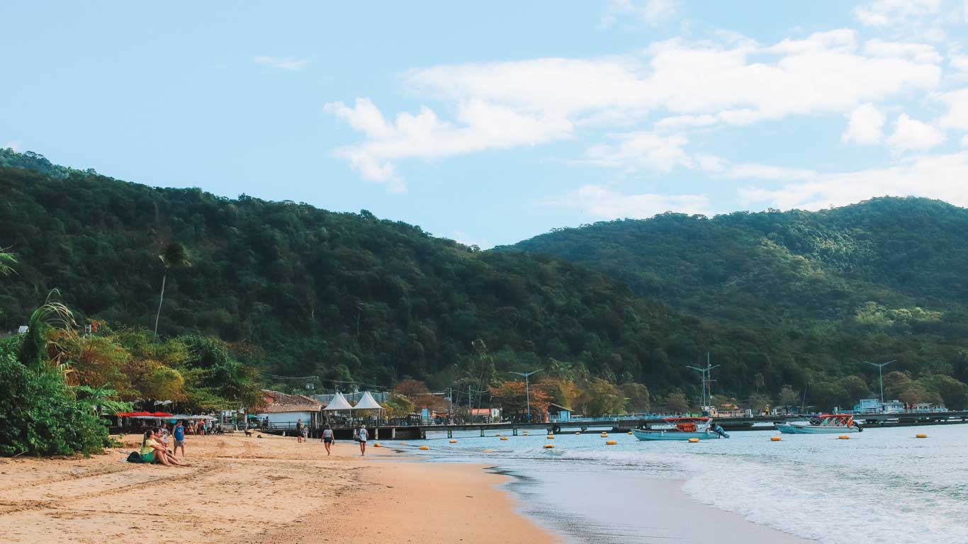 The picturesque beach in Ilha Grande, Rio de Janeiro, with golden sand curving alongside a calm shoreline. Visitors can be seen relaxing on the sand and walking near the water, while green-covered hills rise in the background under a bright blue sky.