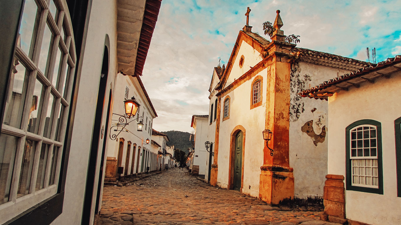 A cobblestone street in the Historic Center, the best place to stay in Paraty, at sunset, with lanterns glowing, illuminating the façade of an old church and colonial buildings. The peaceful and picturesque atmosphere highlights the beauty of the historic architecture.