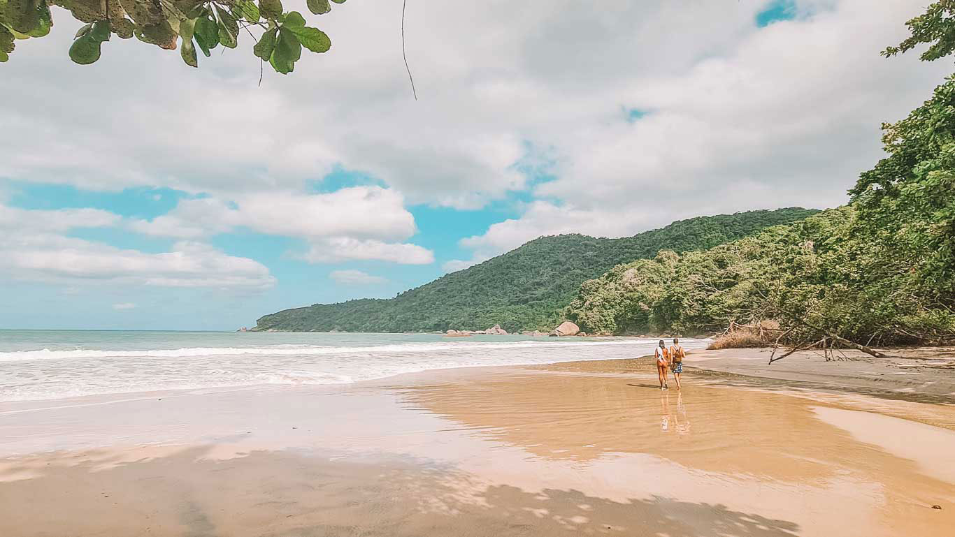 Cachadaço Beach in Trindade, Paraty, with two people walking along the shore under a partly cloudy sky. The beach is surrounded by lush vegetation and hills, creating a peaceful and natural setting.