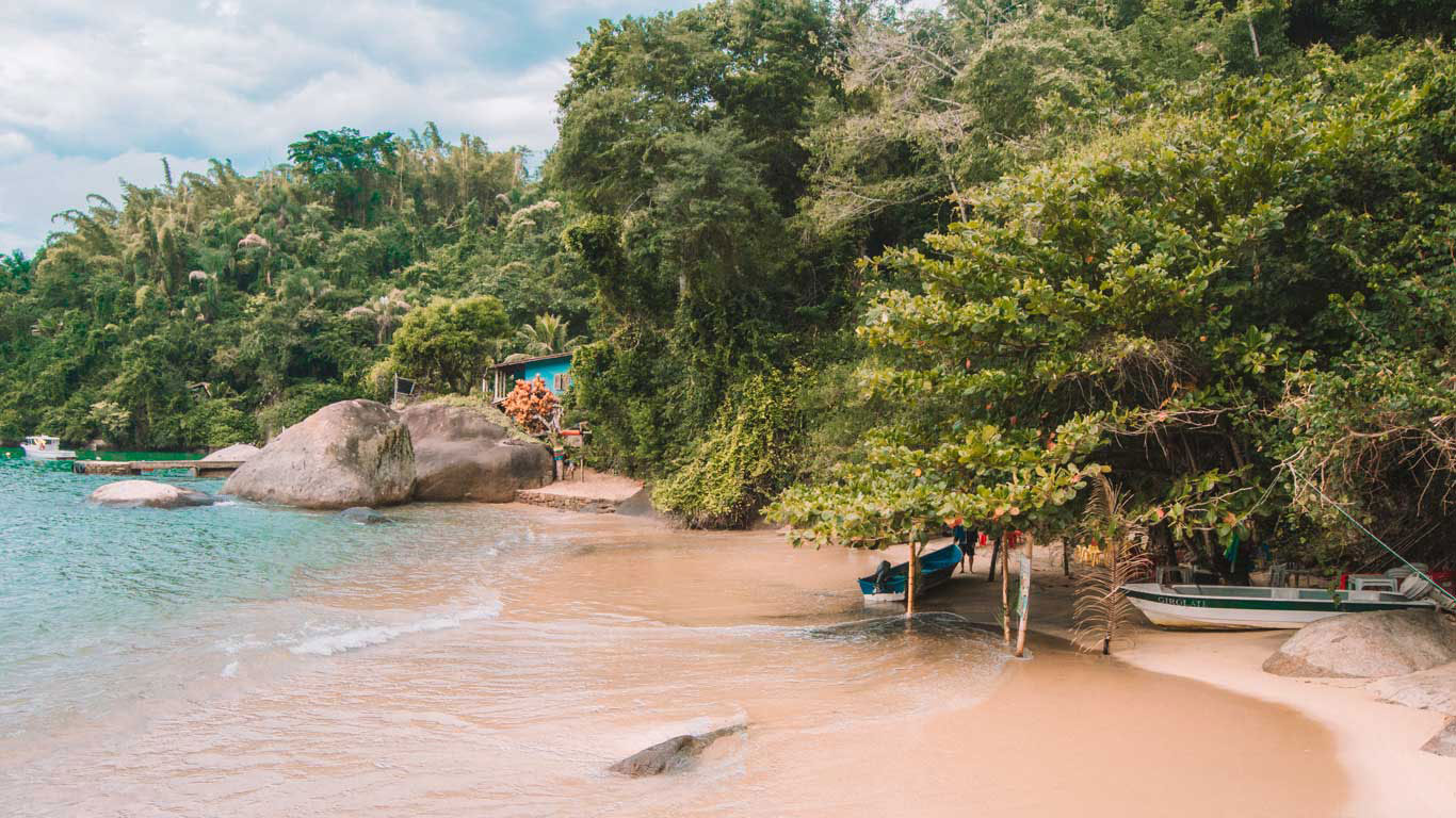 A small beach in Paraty, Rio de Janeiro, Brazil, with crystal-clear waters and dense surrounding vegetation. A blue house and several small boats near the shore create a cozy and serene atmosphere. Large rocks are scattered along the sand and water, adding to the picturesque natural landscape.