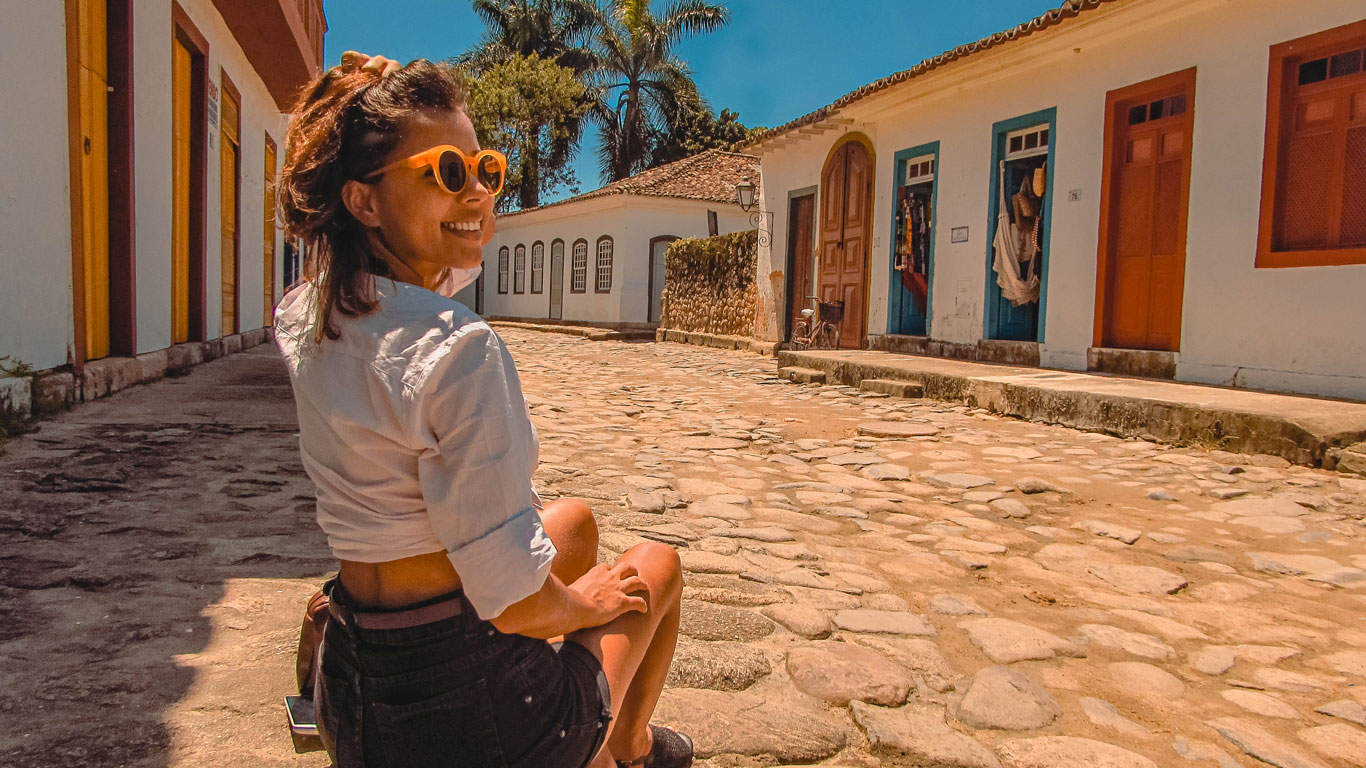 A woman wearing a white shirt and orange sunglasses is sitting on a cobblestone street in the historic center of Paraty, Rio de Janeiro, smiling and enjoying a sunny day. The street is lined with colorful colonial buildings, with doors and windows painted in vibrant tones.